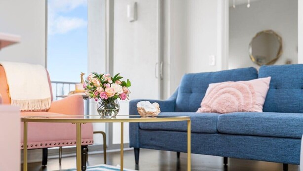 Una sala de estar moderna con un sofá azul, un sillón rosa y una mesita con un jarrón de flores.