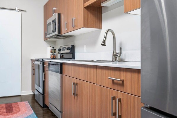 A modern kitchen with wooden cabinets, stainless steel appliances, and a white countertop. - 2BR TC Stay I Near DT + Trails (Traverse City)