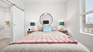 A bedroom with a large bed, a round mirror above it, two bedside tables with lamps, and a window with blinds. - Stylish 2BR Suite Near DT Traverse City + Smart TV (Traverse City)