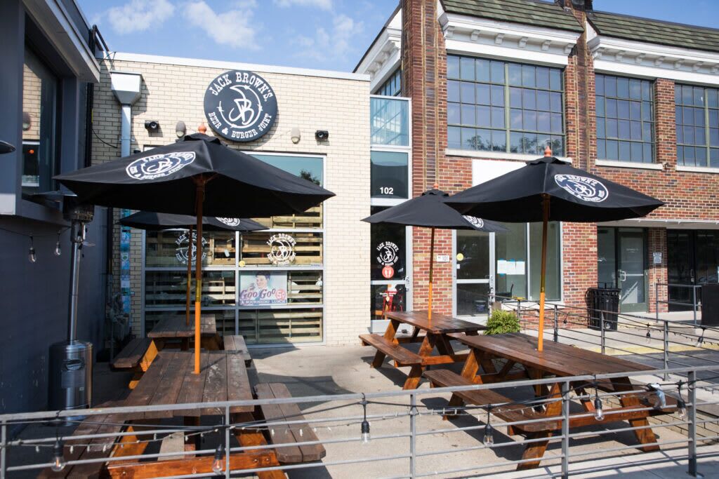 Un espace extérieur aménagé avec des tables en bois et des parasols noirs, un bâtiment en briques sur lequel on peut lire l’inscription « Jack Brown’s Brewery & Bar », et un ciel dégagé.