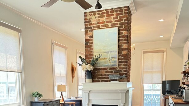 A spacious room with a fireplace, wooden flooring, a ceiling fan, and a large window. - The Kelley House | Suite 2 - 1st floor (Pensacola)