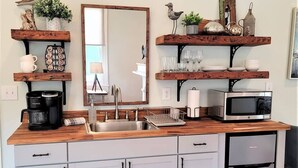 A kitchen with a wooden countertop, a sink, a coffee maker, a microwave, and a dishwasher. There are shelves with decorative items and a mirror above the sink. - The Kelley House | Suite 2 - 1st floor (Pensacola)