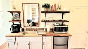 A modern kitchen with white cabinets, a stainless steel dishwasher, a microwave, and wooden countertops. There are shelves with decorative items and a large mirror above the sink. - The Kelley House | Suite 3 w/Balcony (Pensacola)