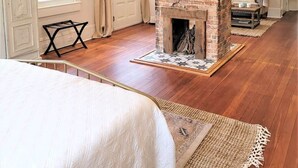 Une chambre avec une cheminée en briques, un plancher en bois, un lit avec une literie blanche et un ventilateur de plafond.