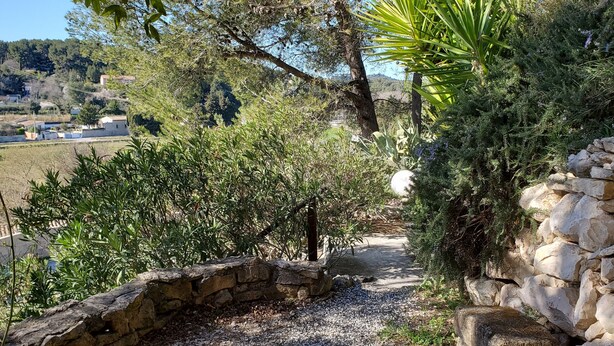 Uma trilha de pedra serpenteia por um jardim com vegetação exuberante e um muro de pedra em um dos lados.