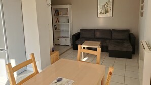 Uma sala de estar com um sofá cinza escuro, uma estante branca, uma pequena mesa de madeira e uma geladeira branca.