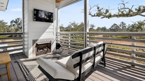 A covered outdoor seating area with a fireplace, a bench, and a TV mounted on the wall. - Heated Pool, Steps to Beach, Stylish Interior, Amenities Galore!  (Waveland)