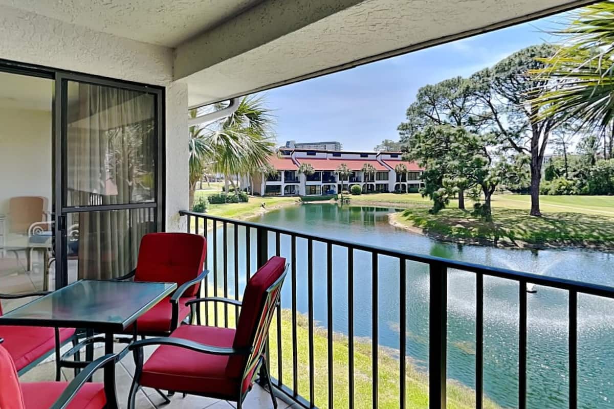 A balcony with a glass table and red chairs, overlooking a body of water and a building with a red roof.