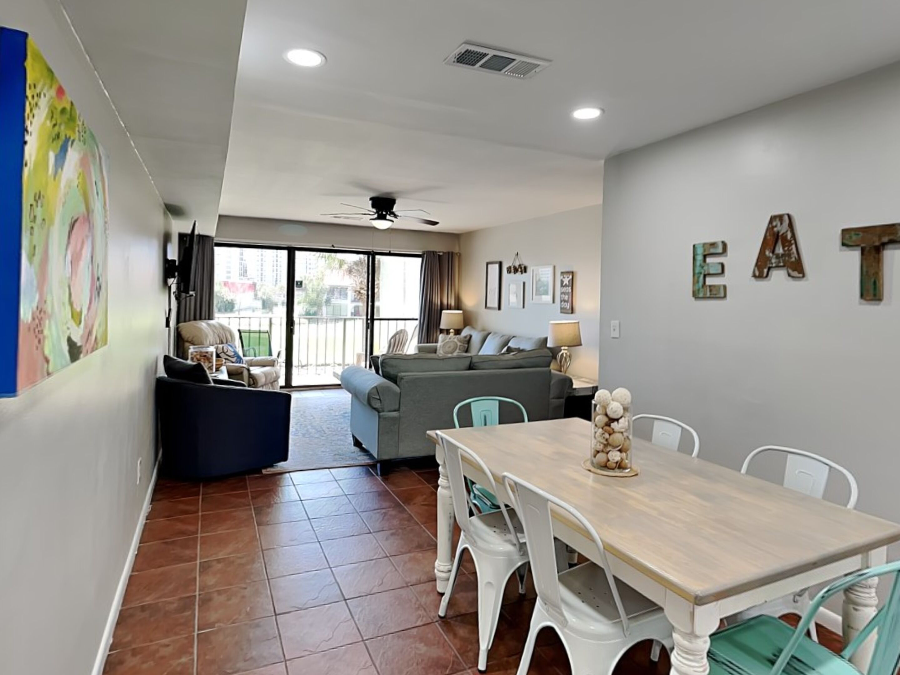 A modern living room with a sofa, dining table, and chairs. There is a ceiling fan, a large window with curtains, and a wall with the word "EAT" in rustic letters.