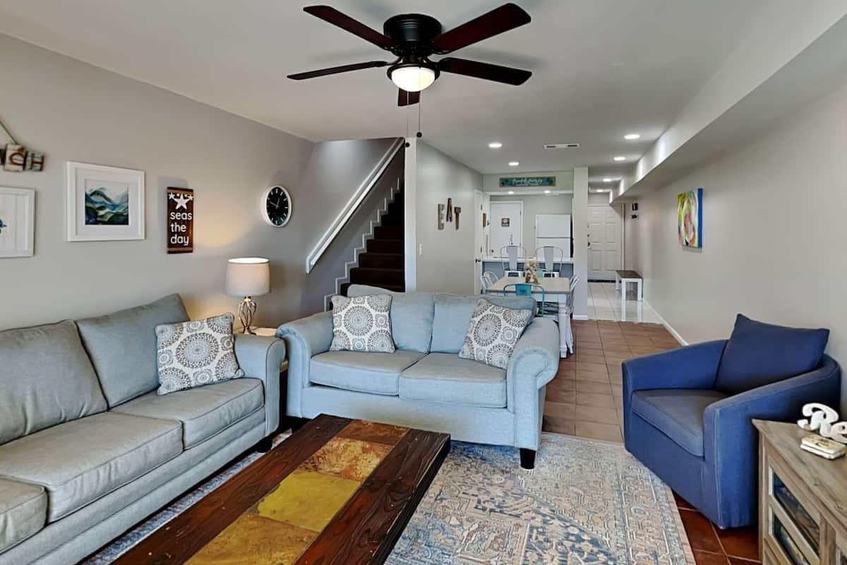 A spacious living room with a grey sofa, a blue armchair, and a patterned rug. A staircase leads to a dining area with a white table and chairs.