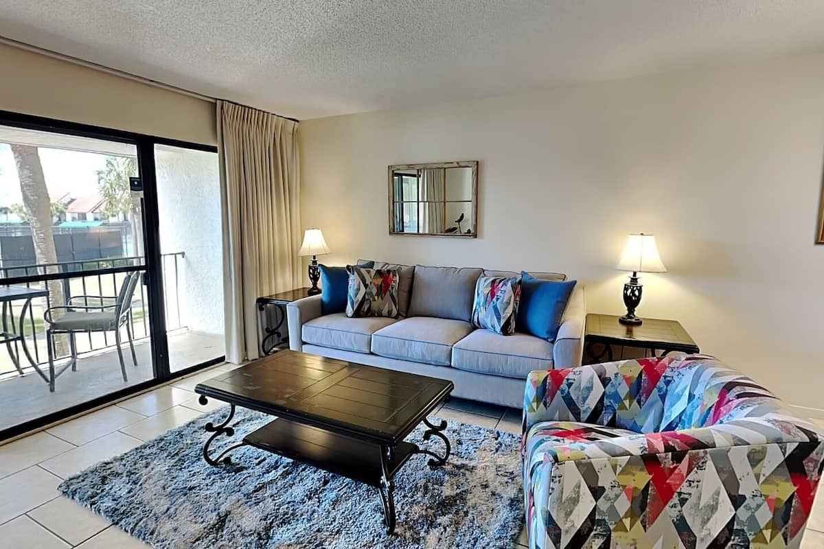 A living room with a sofa, a coffee table, and a colorful armchair. There is a balcony with chairs and a table outside.