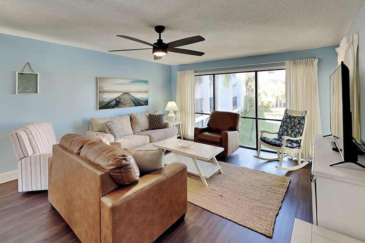 A living room with a brown leather sofa, a white coffee table, a brown armchair, and a rocking chair. There is a ceiling fan, a framed picture on the wall, and a large window with curtains.