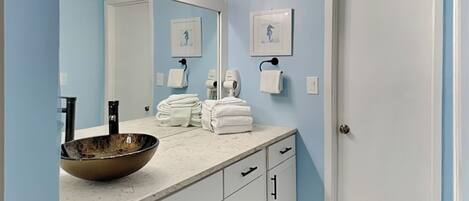 A bathroom with a large mirror, a sink with a bowl, towels stacked on the counter, and a door.
