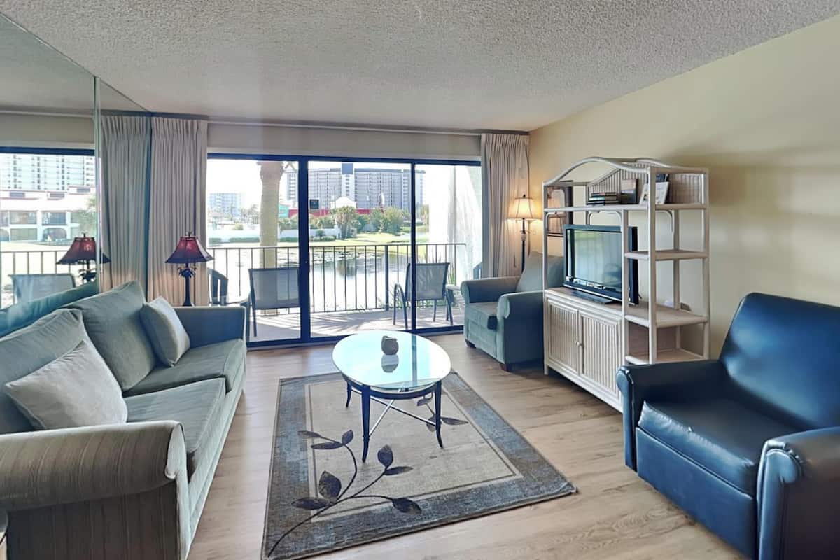 A modern living room with a sofa, armchair, TV stand, and a balcony with a view of the city.