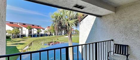 A balcony with a view of a pond, a bench, and a building in the background.