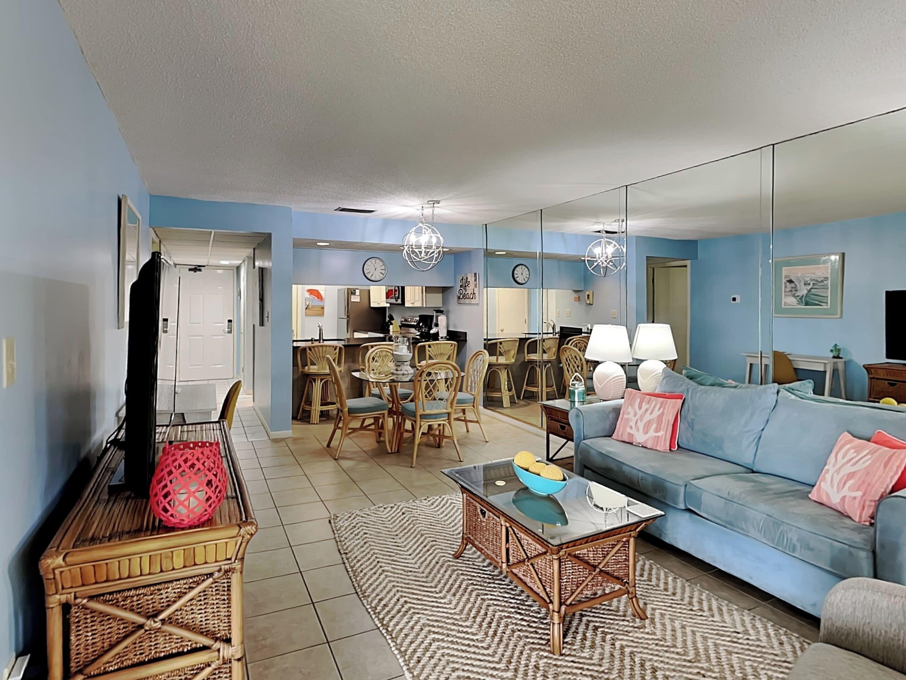 A living room with a blue sofa, wicker chairs, a glass coffee table, and a dining area with wicker chairs and a table.