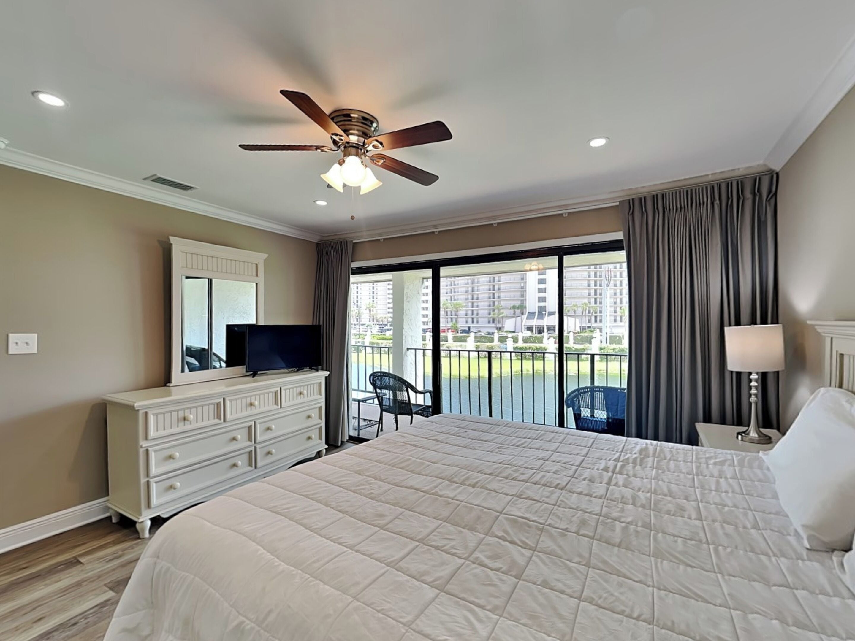 A hotel room with a large bed, a ceiling fan, a television on a dresser, and a balcony with a view of the city.