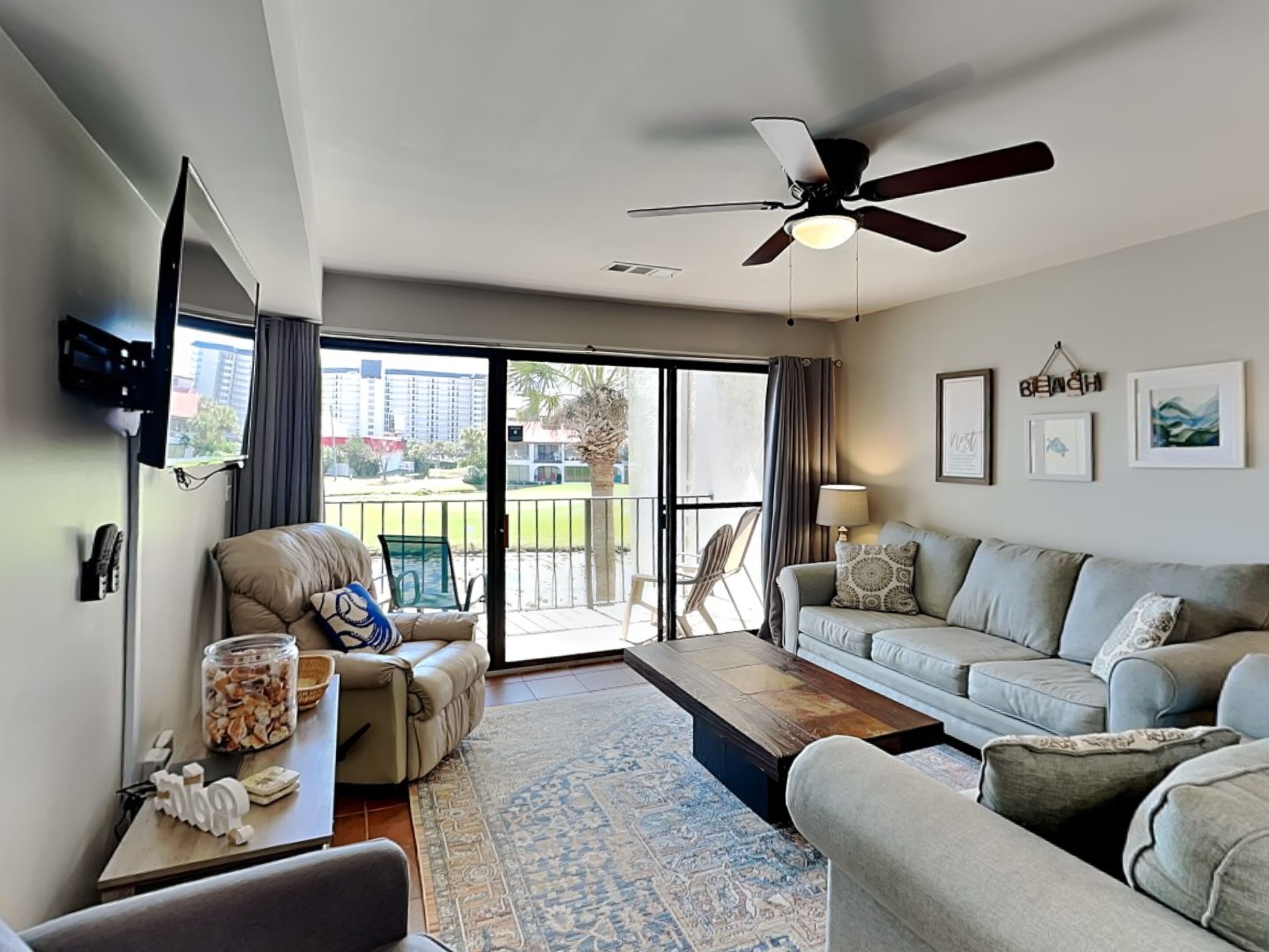 A living room with a sofa, a coffee table, and a TV mounted on the wall. There is a balcony with a view of buildings and a palm tree.