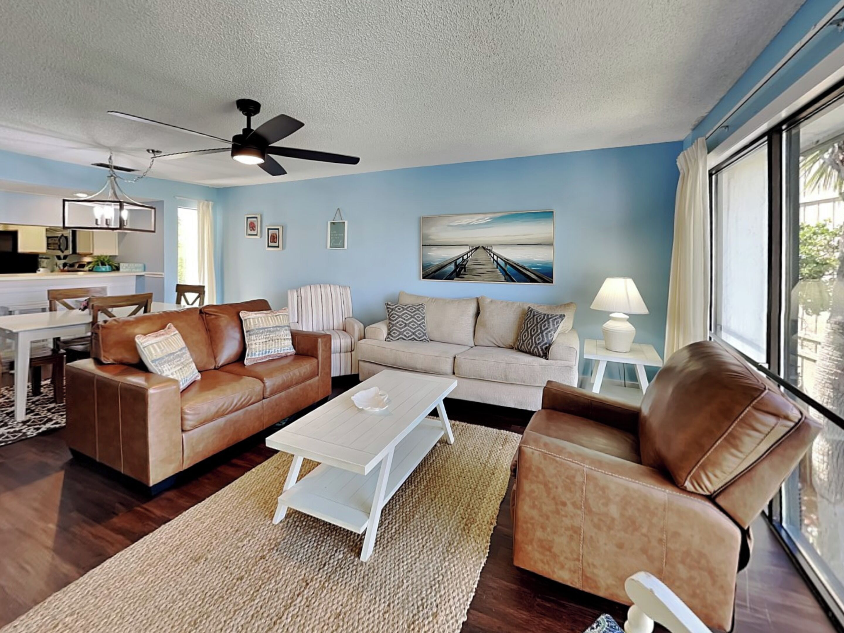 A living room with a brown leather sofa, a beige sofa, and a white coffee table. There is a ceiling fan, a painting on the wall, and a window with a view of the outside.