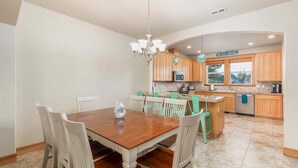 A dining area with a wooden table and white chairs, a kitchen with wooden cabinets and a sign reading "BEACH HOUSE", and a chandelier. - Sacred Waters Retreat -updated townhome, near beach. Dog OK. (Pacific City)