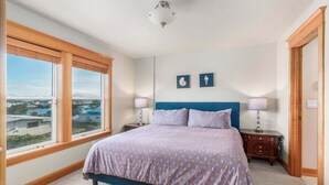 A bedroom with a large window, a bed with a blue headboard, two bedside tables with lamps, and two framed pictures on the wall. - Sacred Waters Retreat -updated townhome, near beach. Dog OK. (Pacific City)