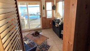 Un intérieur en bois avec une grande fenêtre donnant sur un plan d’eau, un coin repas avec une table et des chaises, et un espace cuisine équipé d’un réfrigérateur et de placards.