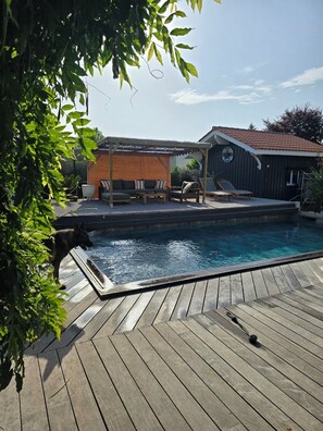 Um deck de madeira com piscina, um espaço coberto de varanda e um pequeno prédio com um relógio.