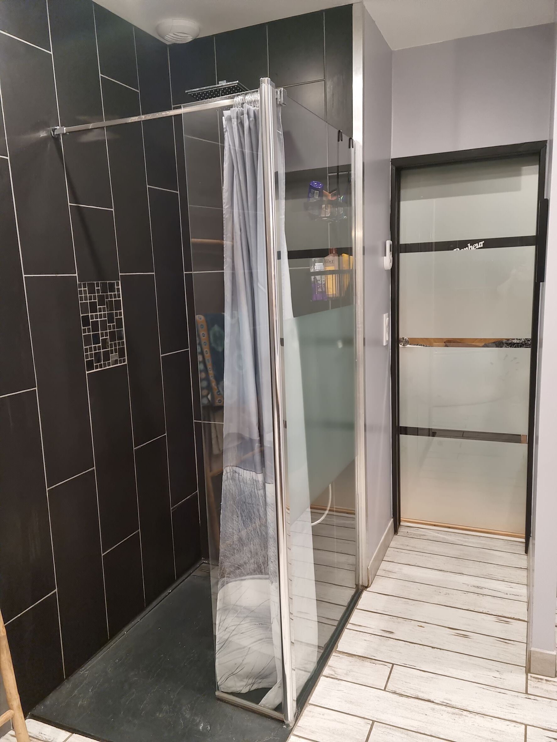 A modern bathroom with a glass shower enclosure, dark tiled walls, and a white tiled floor.