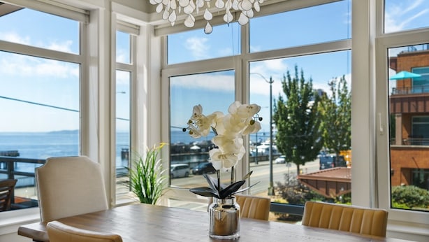 A dining area with a table and chairs, a vase with white flowers, and large windows offering a view of the sea and buildings. - 🚢Luxury Seattle Waterfront Walk to Cruise Ship & Pike Place -Waterfront Views🎡 (Seattle)