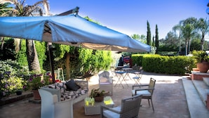 Um pátio externo com mobiliário de vime, uma mesa e um grande guarda-sol.