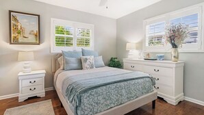 Ein Schlafzimmer mit Bett, Nachttischen, einem Schminktisch, einem Deckenventilator und einem Fenster mit Fensterläden.