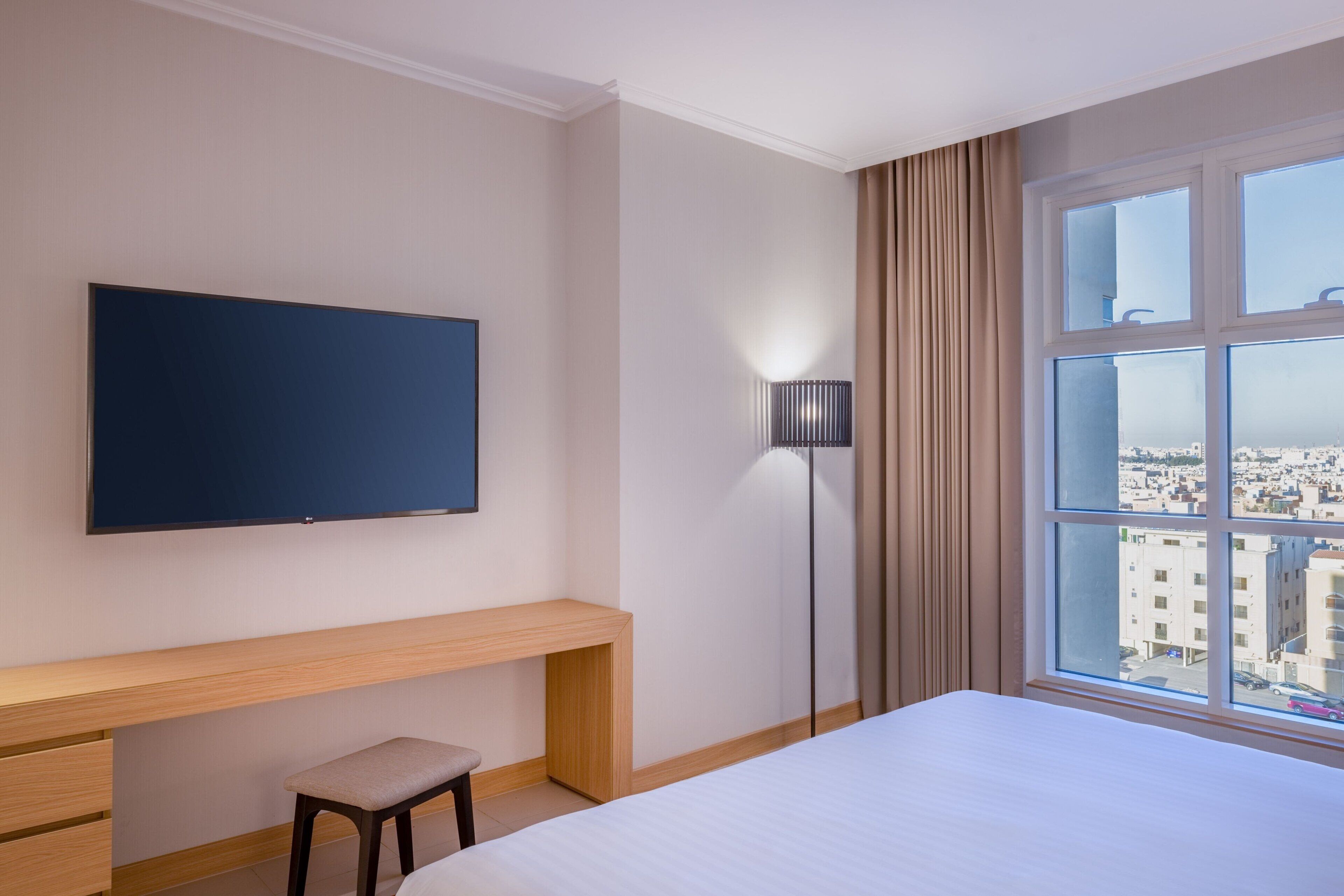 A hotel room with a bed, a TV mounted on the wall, a desk with a chair, a floor lamp, and a window with a city view.