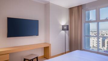 A hotel room with a bed, a TV mounted on the wall, a desk with a chair, a floor lamp, and a window with a city view.