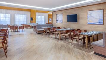 A spacious dining area with multiple tables and chairs, wooden flooring, and large windows allowing natural light.