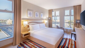 Een hotelkamer met een groot bed, twee nachtkastjes met lampen, een bureau met stoel en een televisie. Er zijn grote ramen met gordijnen en kunst aan de muur.