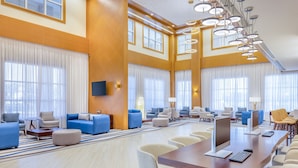 Een ruime hotel lobby met verschillende zithoeken, een centrale tafel met computers en grote ramen voorzien van gordijnen.