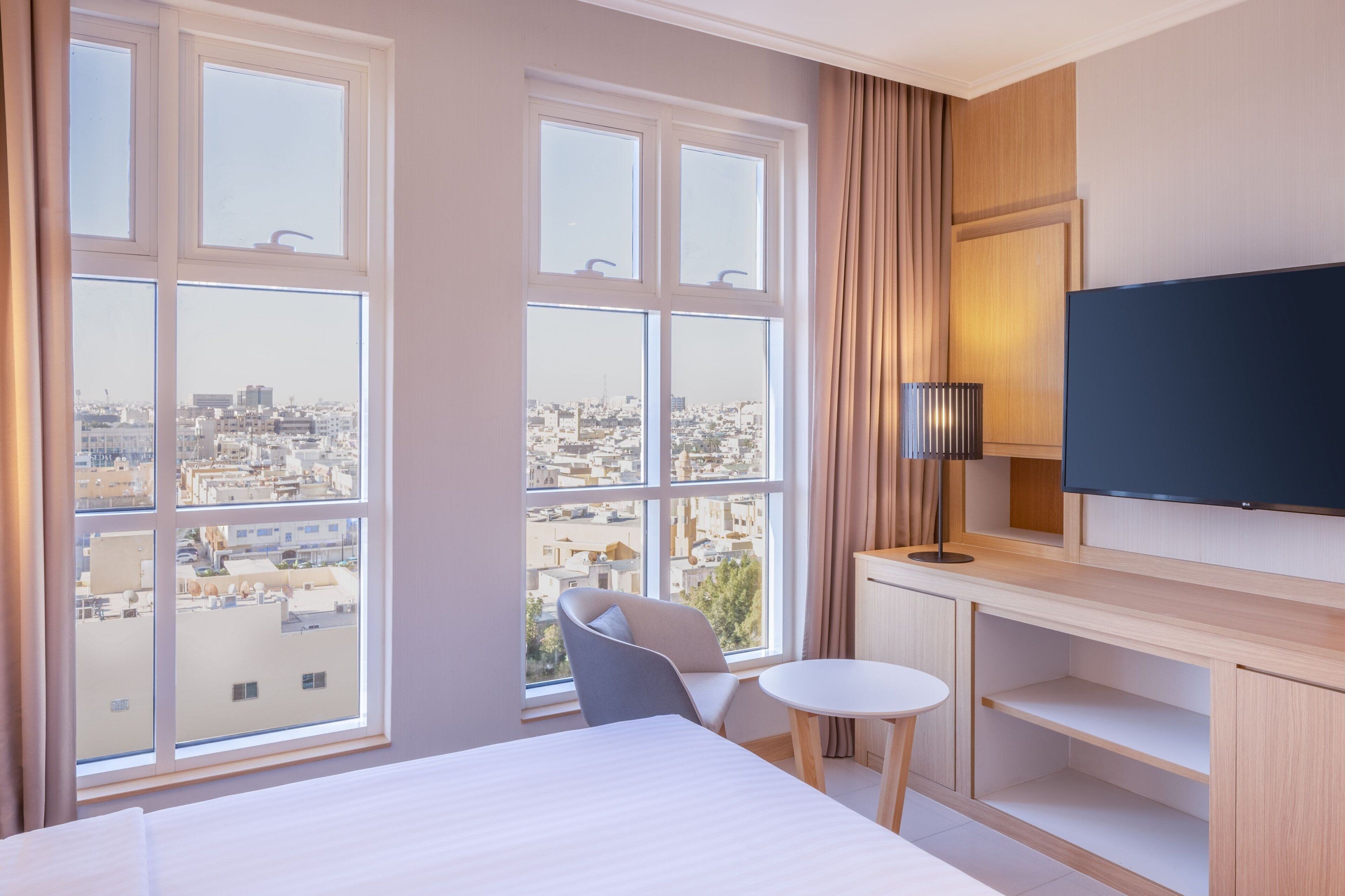 A hotel room with a large window, a bed, a chair, a small table, a TV, and a cabinet.