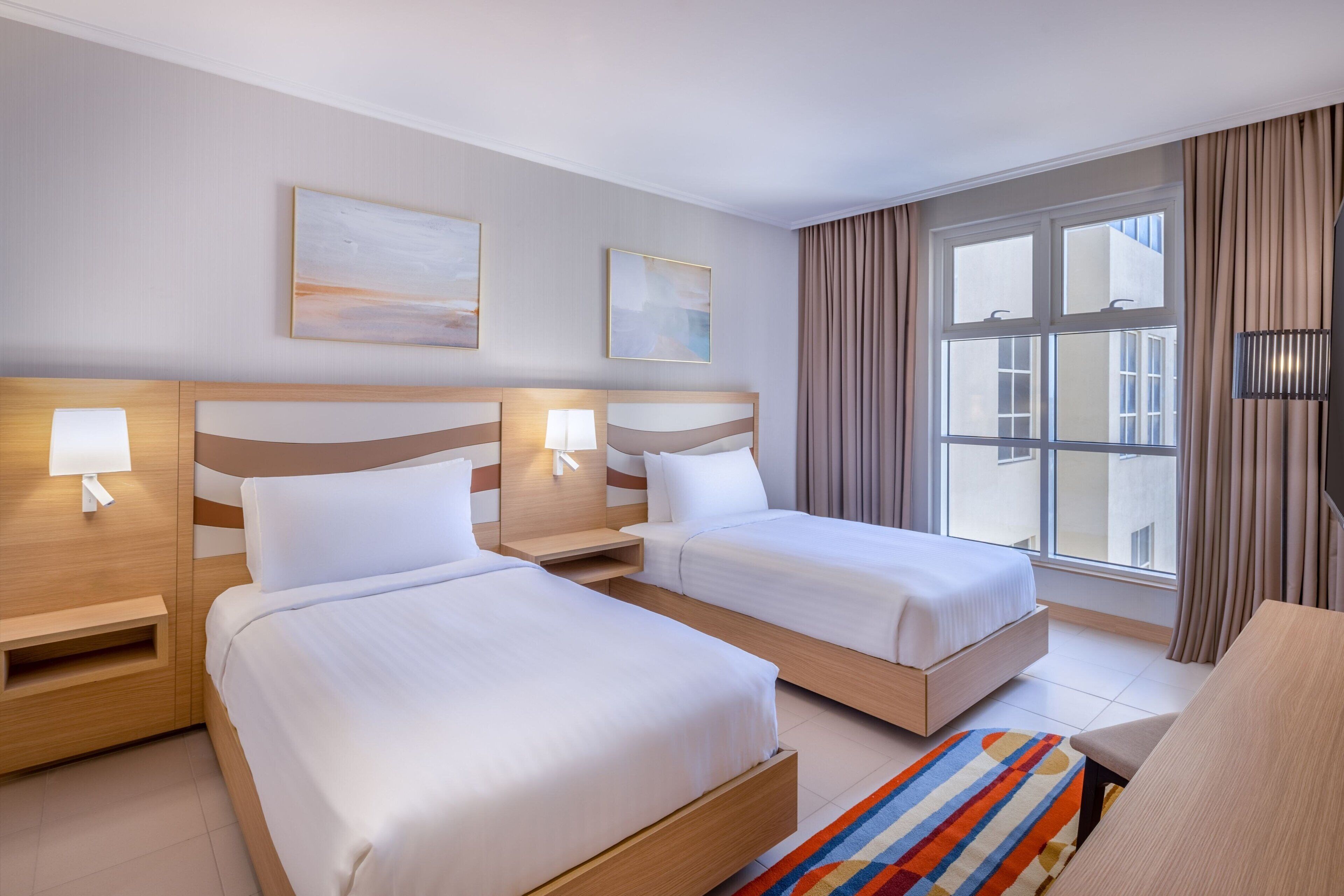 Ein Hotelzimmer mit zwei Betten, einem Schreibtisch, einem Stuhl und einem großen Fenster mit Vorhängen.