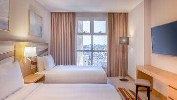 A hotel room with two beds, a desk, a TV, and a view of the city.