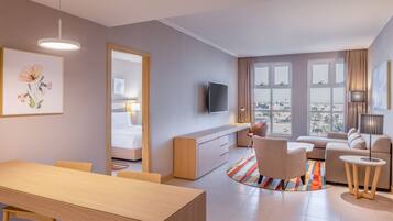 A modern hotel room with a dining area, a sofa, a TV, and large windows offering a city view.