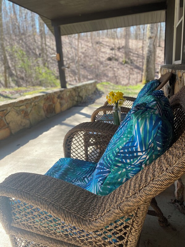 A wicker chair with a blue patterned cushion, a small table with a vase of yellow flowers, and a stone wall in a covered outdoor area.