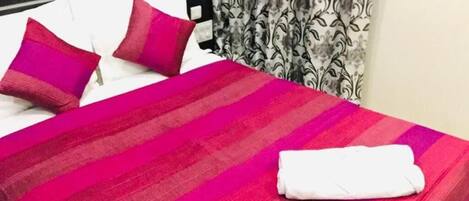 Un lit bien fait, avec une couvre-lit à rayures et deux oreillers, une serviette blanche pliée, et un rideau à motifs en arrière-plan.