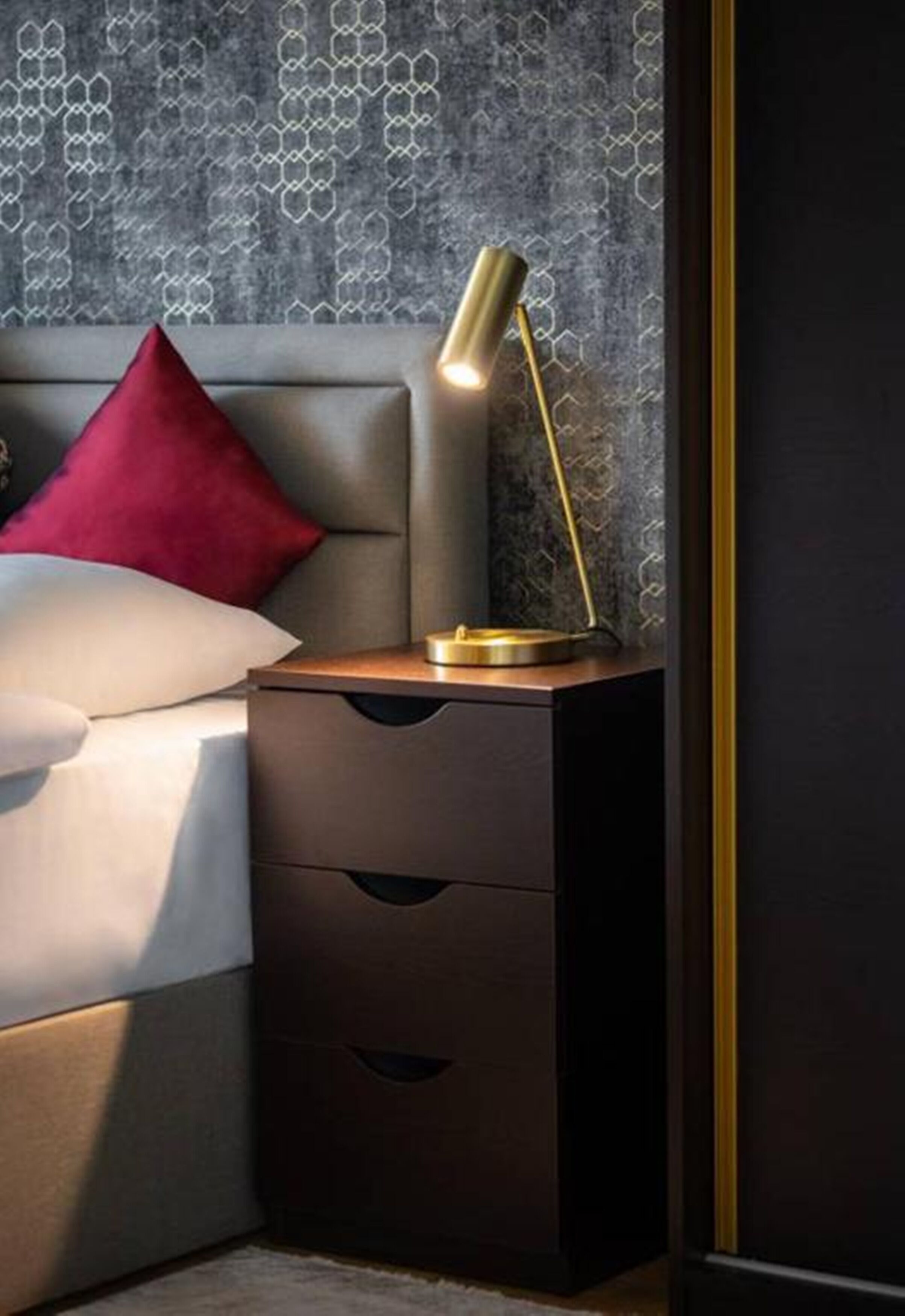 A neatly made bed with a red pillow, a bedside table with a lamp, and a patterned wallpaper.