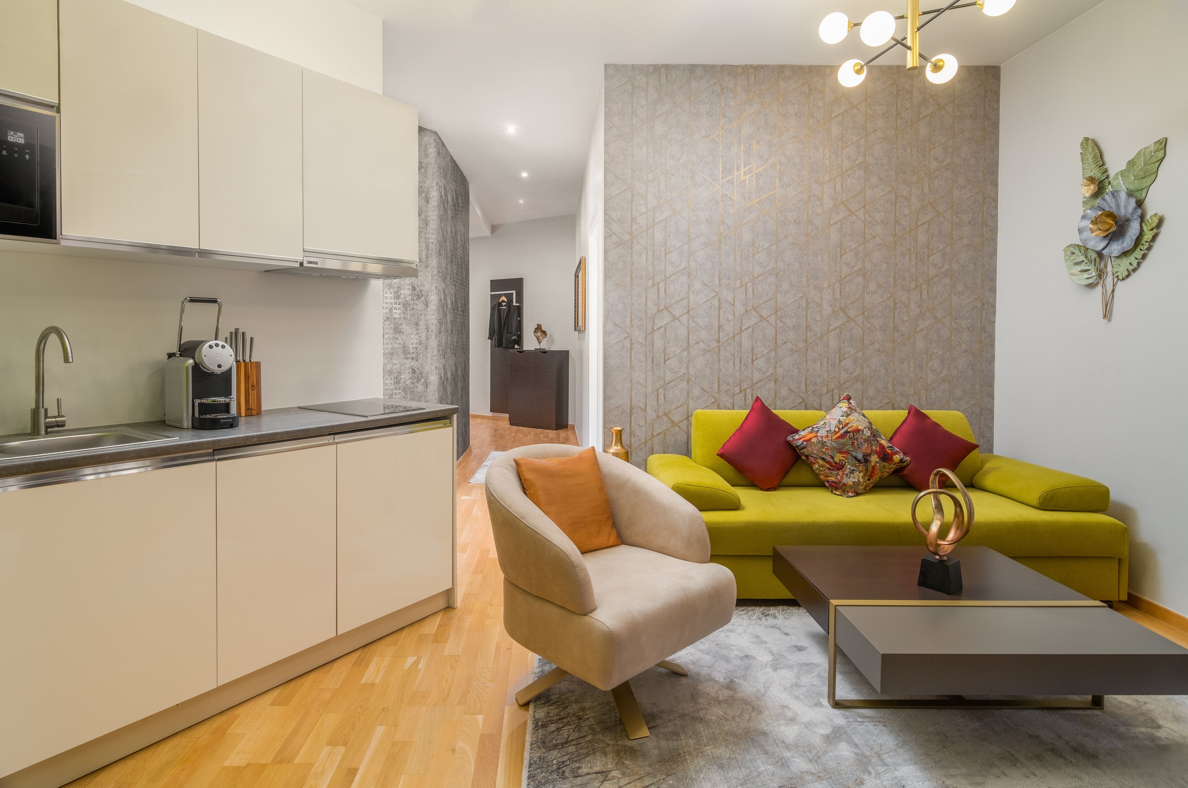 Una cocina moderna con armarios blancos, horno empotrado y cafetera. Hay un sillón color beige y un sofá verde con cojines coloridos en una sala de estar con pared de diseño tipo mármol y una planta decorativa.