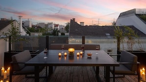 Terraza en la azotea con una mesa para dos, rodeada de velas y copas de vino, con vistas a un paisaje urbano al atardecer.