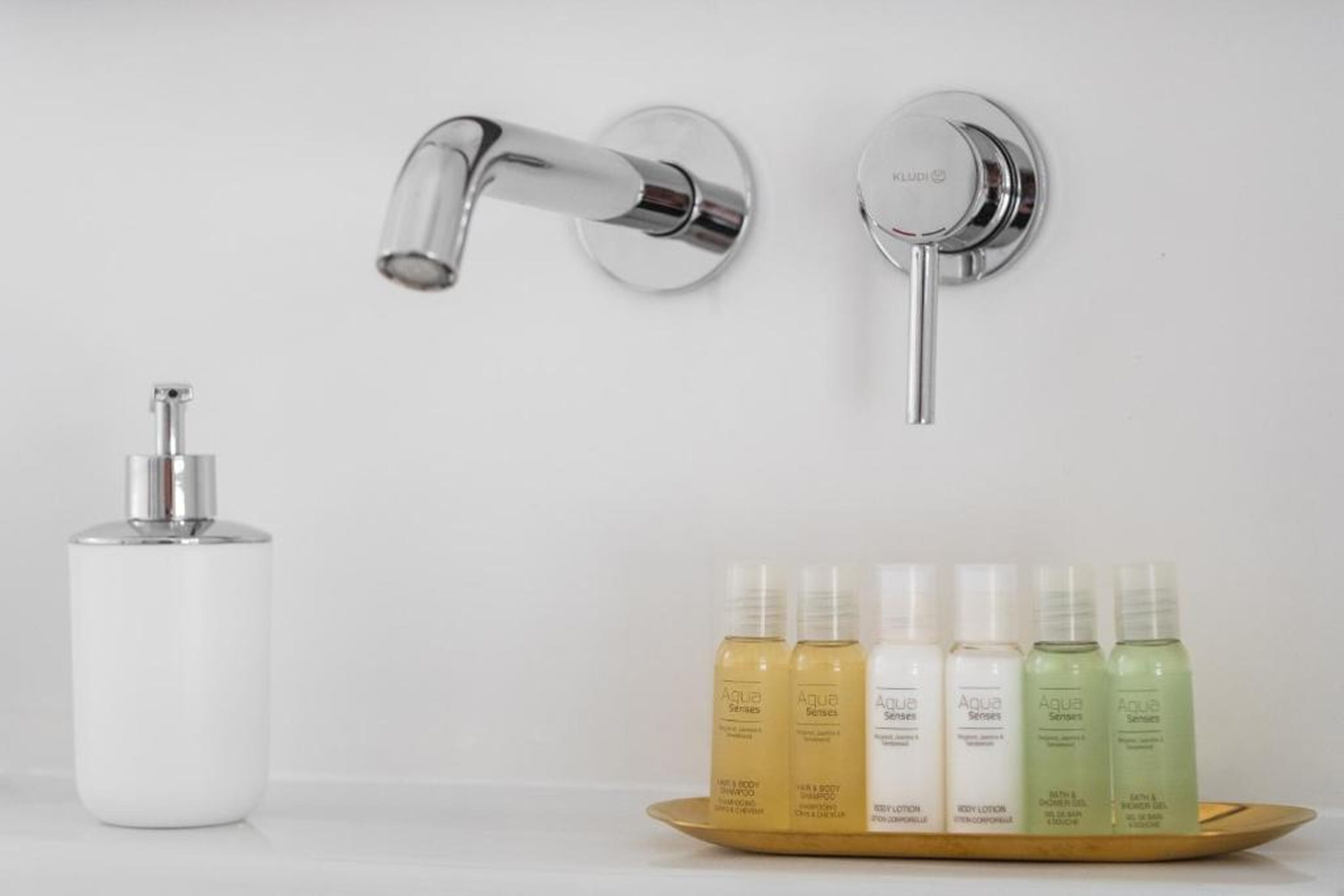 Un baño con un grifo moderno, un dispensador de jabón y una bandeja con cinco botellas de productos de cuidado corporal Aqua Series.