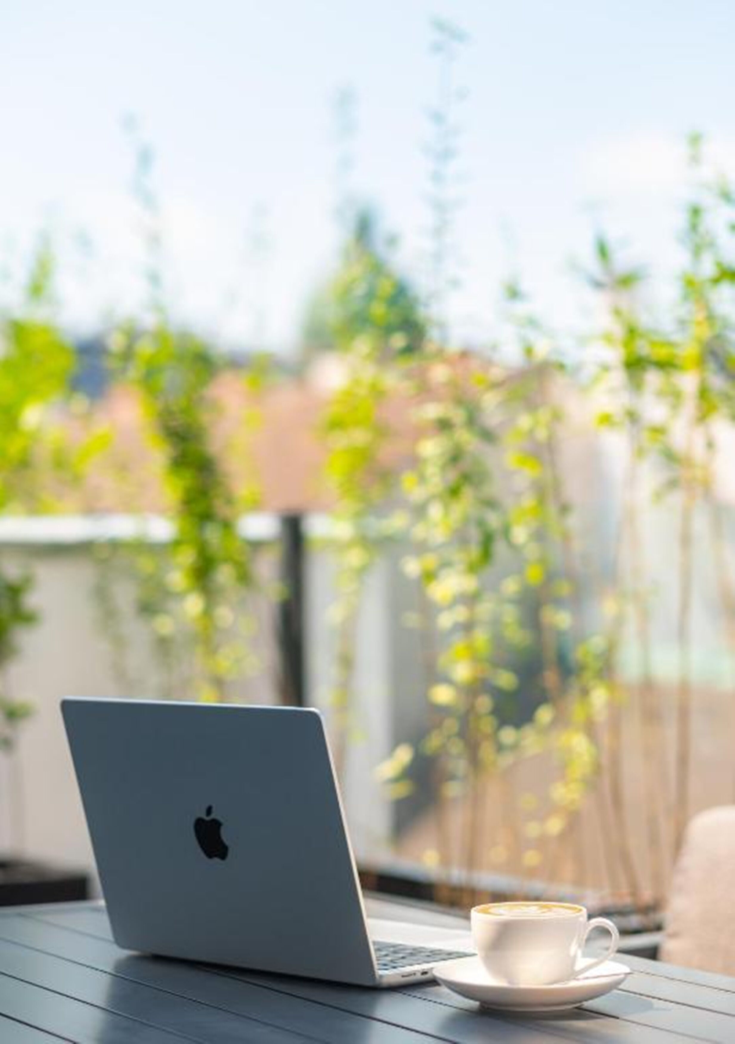 Un ordinateur portable posé sur une table, avec une tasse de café, le tout sur un fond extérieur flou, avec de la verdure.