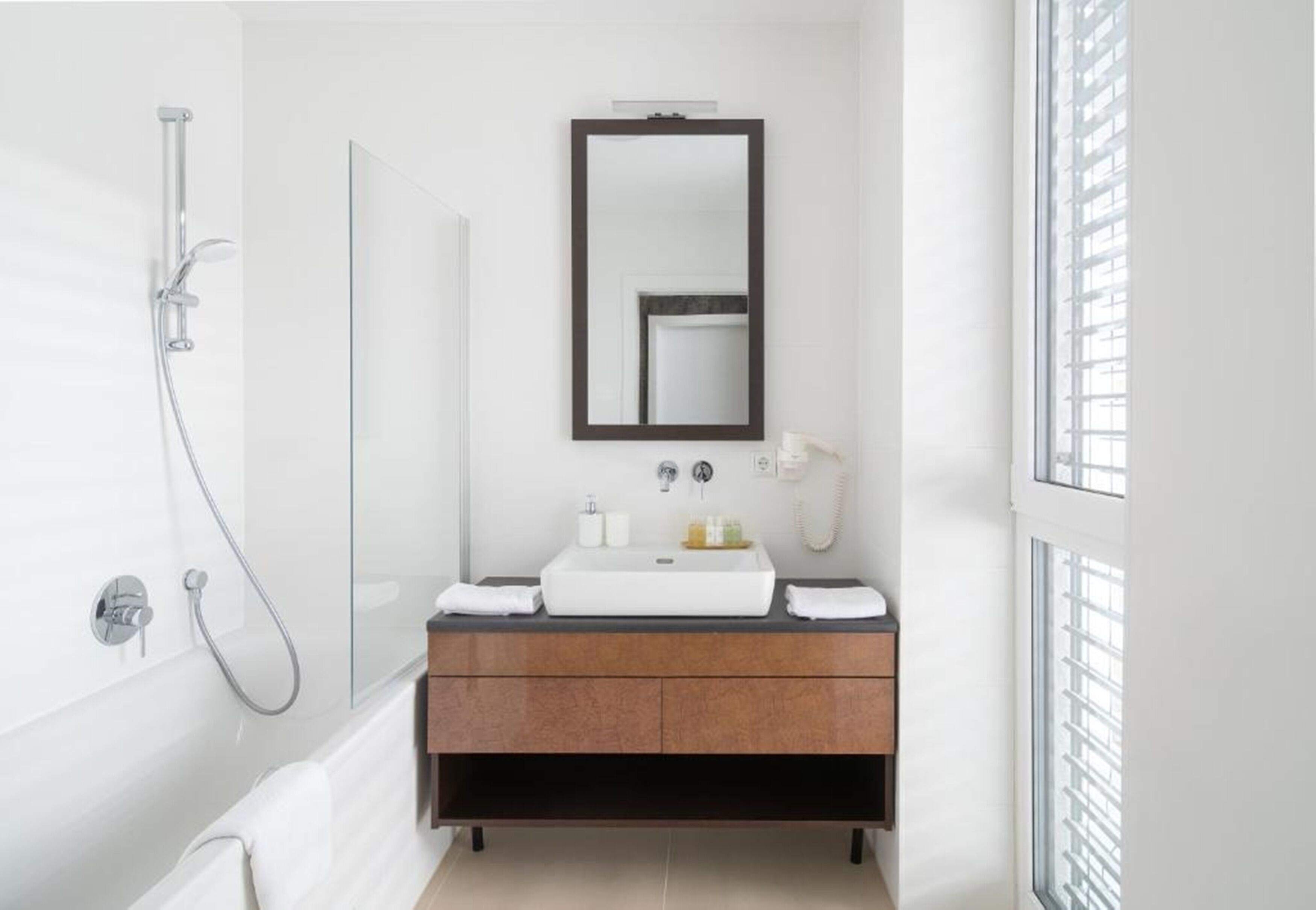 Un baño moderno con ducha de vidrio, un lavamanos blanco y un mueble de tocador de madera oscura, y un espejo grande sobre el mueble.
