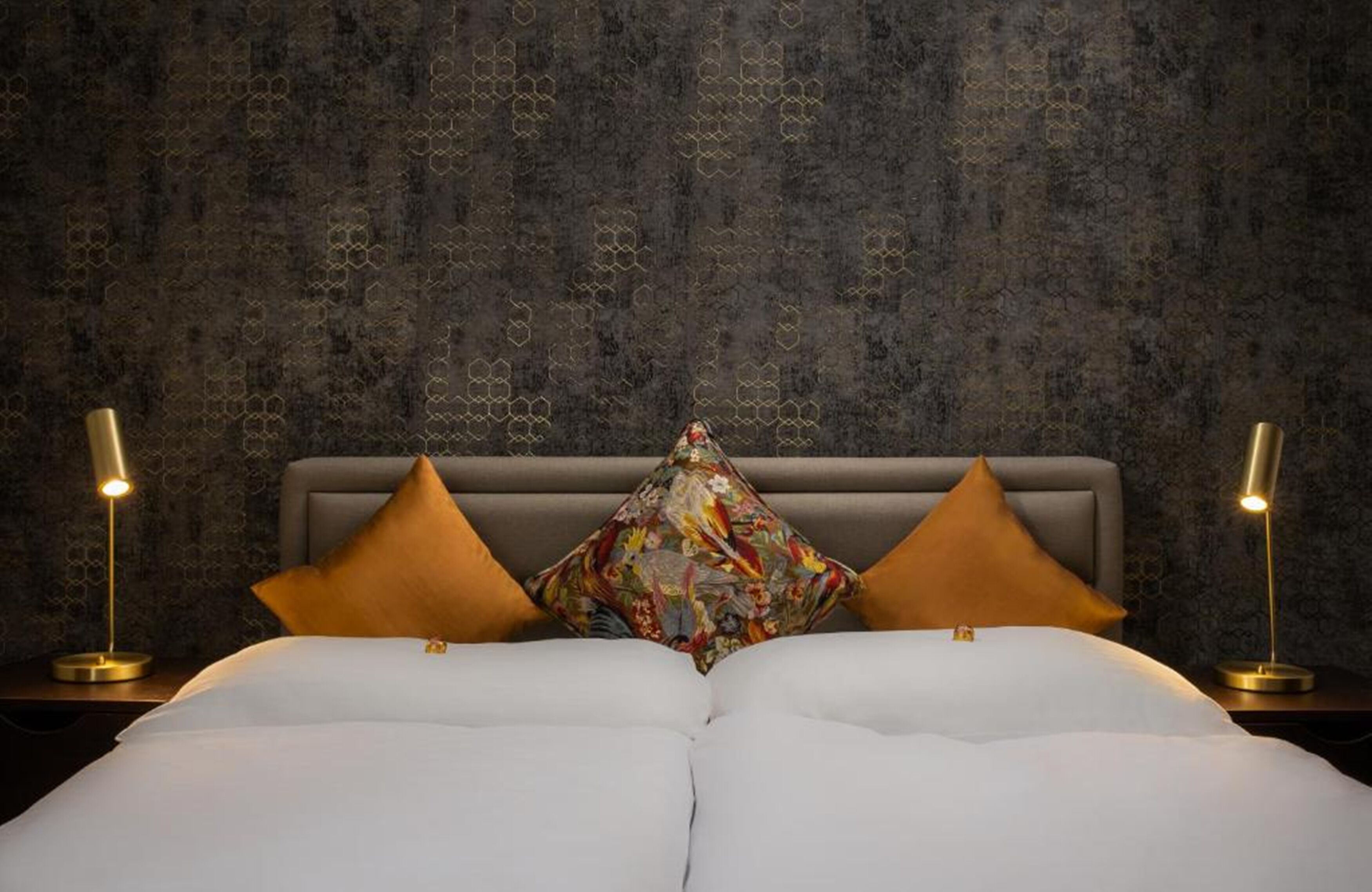 Una cama con sábanas blancas y dos almohadones decorativos, un revestimiento texturizado en la pared de fondo y dos lámparas de noche a cada lado.