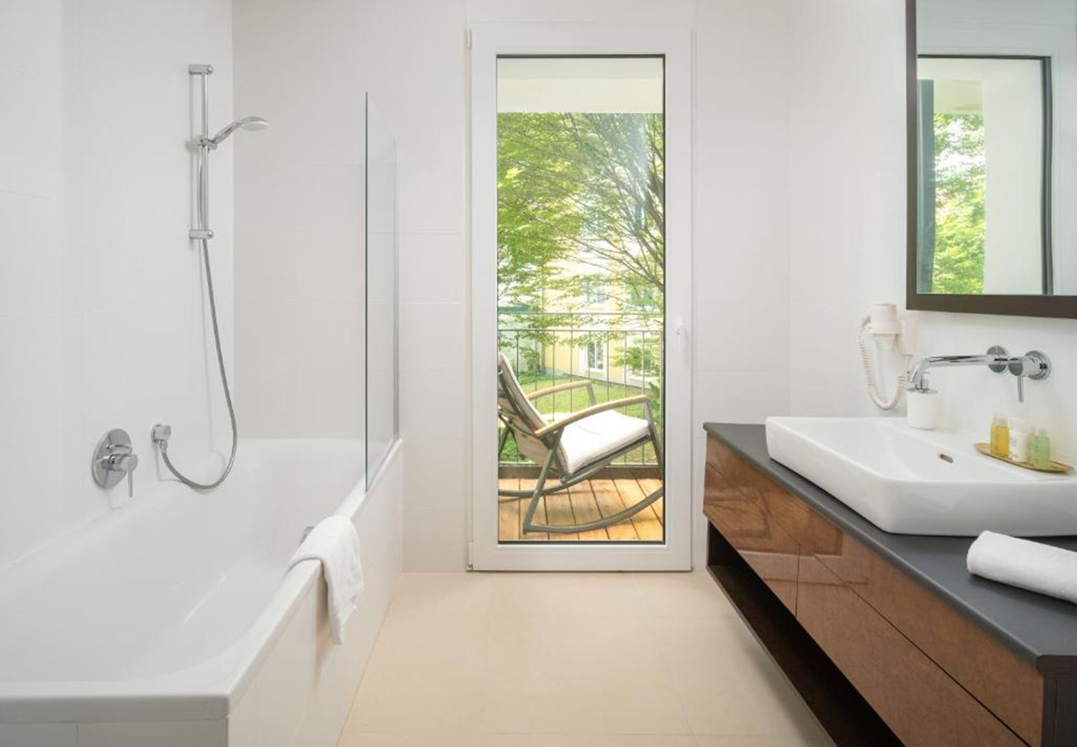 Et moderne badeværelse med badekar, bruser og vask. En glasdør fører ud til en balkon med en stol.